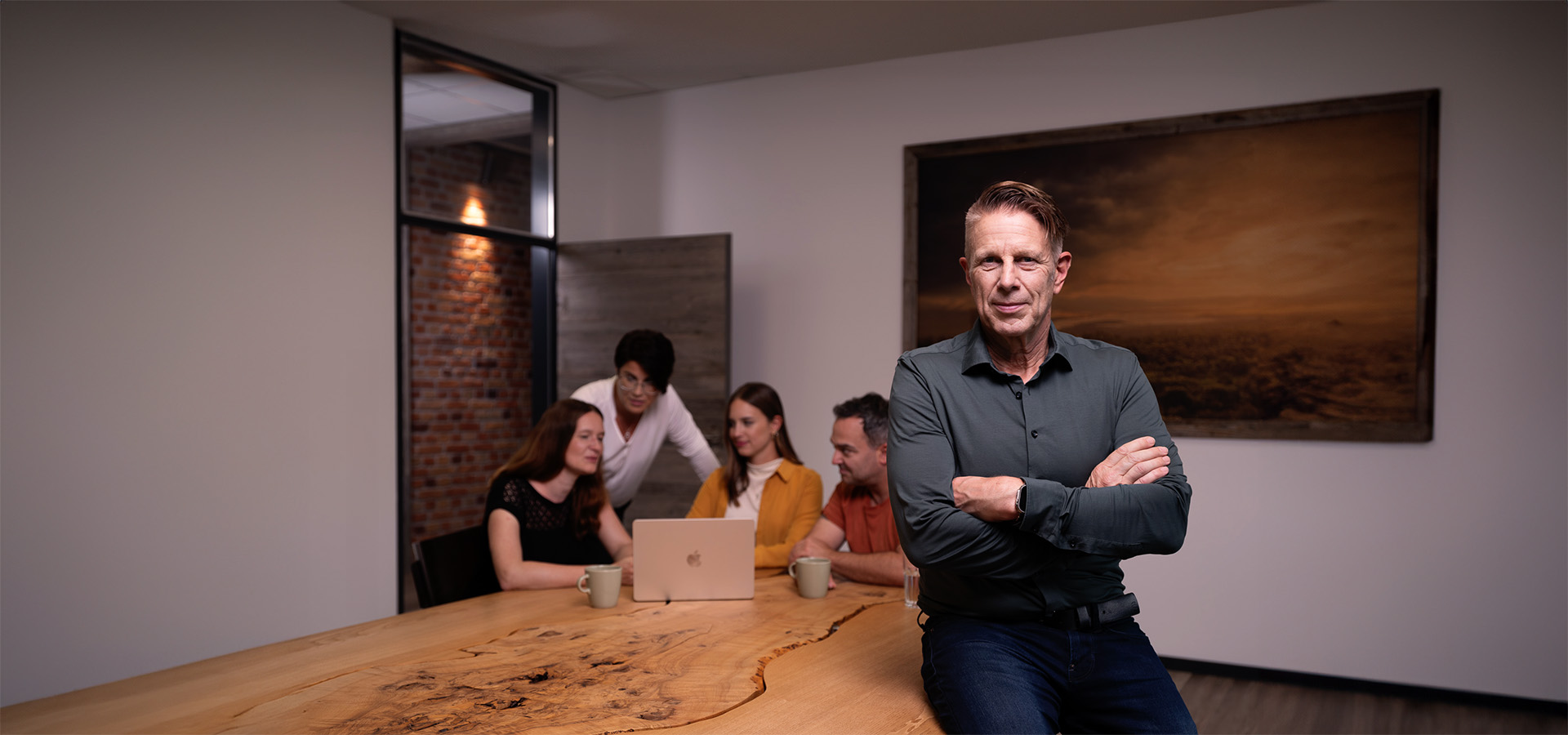 Jochen Greim der Inhaber von Greim Consulting steht mit verschränkten Armen vor einem Tisch, an dem sein Team an einem Laptop arbeitet – ein Bild für die praxisnahe Beratung des Unternehmens in Prozessoptimierung und Supply Chain.