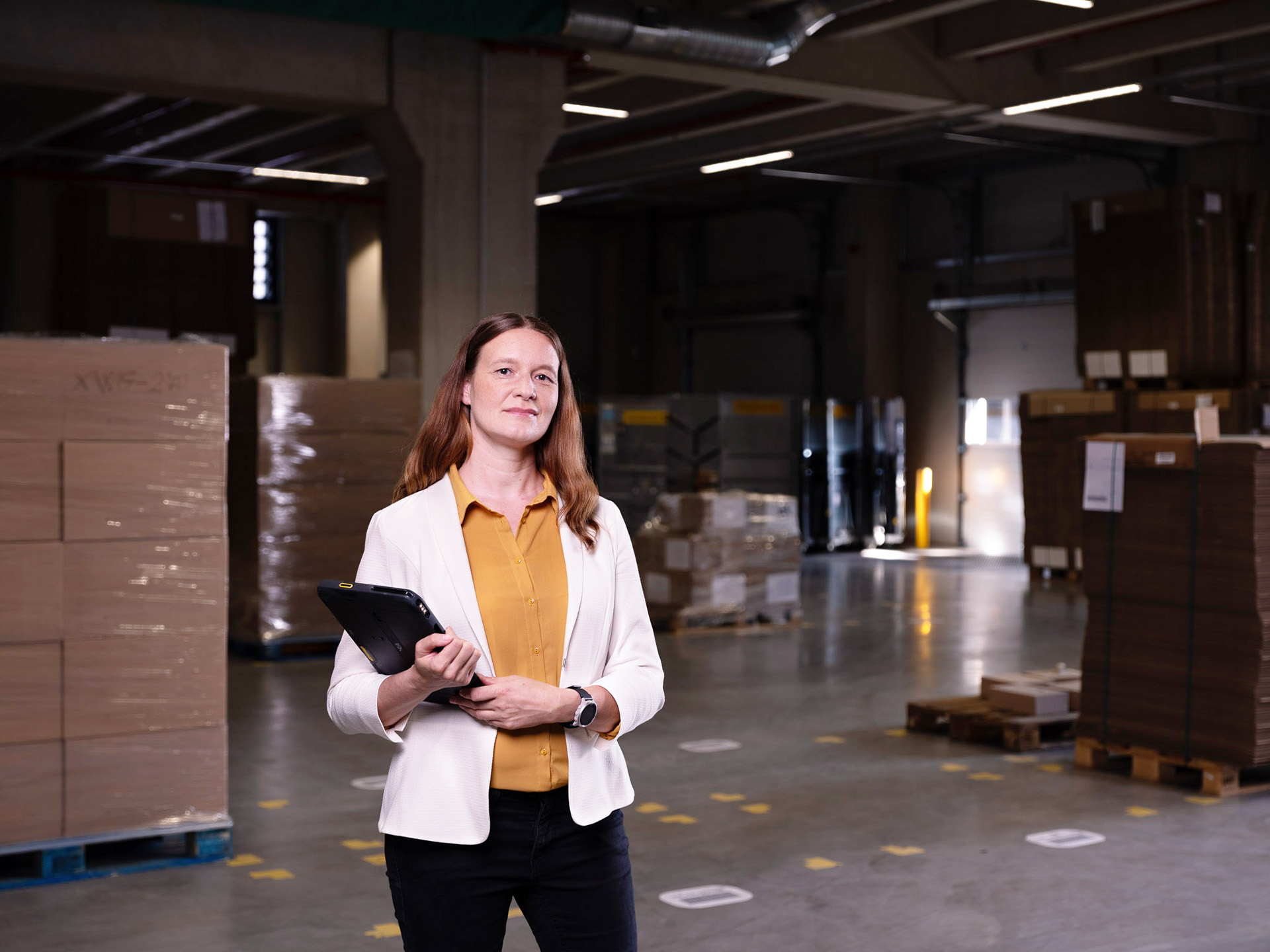 Eine Mitarbeiterin von Greim Consulting, Christina Richter steht in einem Lager mit Paketen und Paletten, hält ein Tablet und repräsentiert die Prozessoptimierung und Supply Chain-Beratung des Unternehmens.