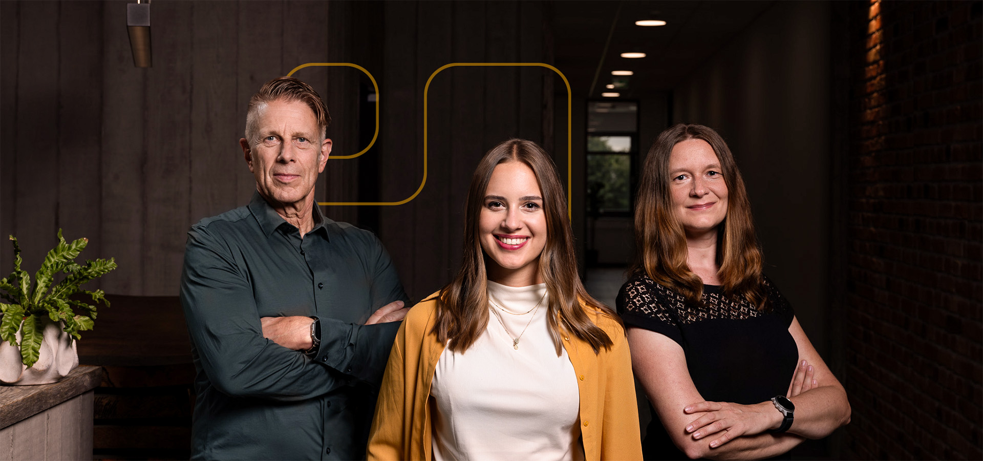 Drei Teammitglieder von Greim Consulting, Jochen Greim, Milena Greim und Christina Richter posieren in einem modernen Büro mit dunklen Wänden und Pflanzendekor, repräsentierend das Beratungsunternehmen mit Fokus auf Prozessoptimierung, Supply Chain und E-Commerce.