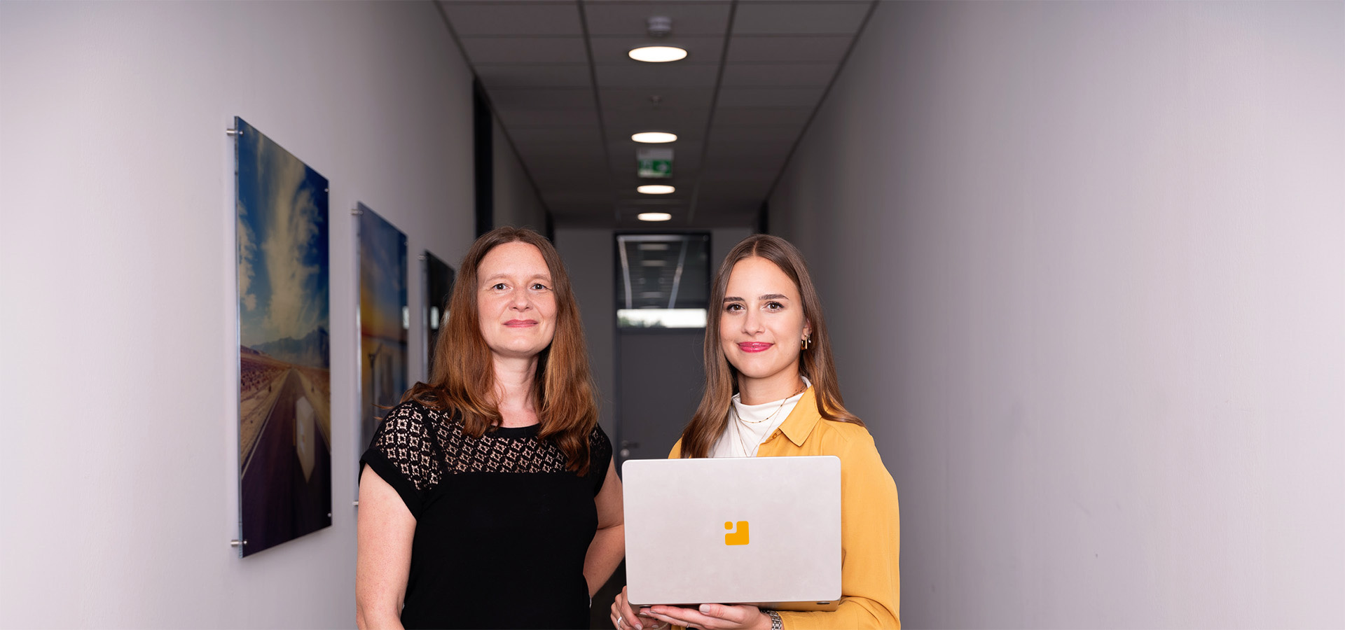 Zwei Frauen, Milena Greim und Christina Richter in einem modernen Büroflur, eine hält ein Laptop mit dem Greim Consulting Logo, repräsentieren das Beratungsunternehmen mit Fokus auf Prozessoptimierung, Supply Chain und E-Commerce.